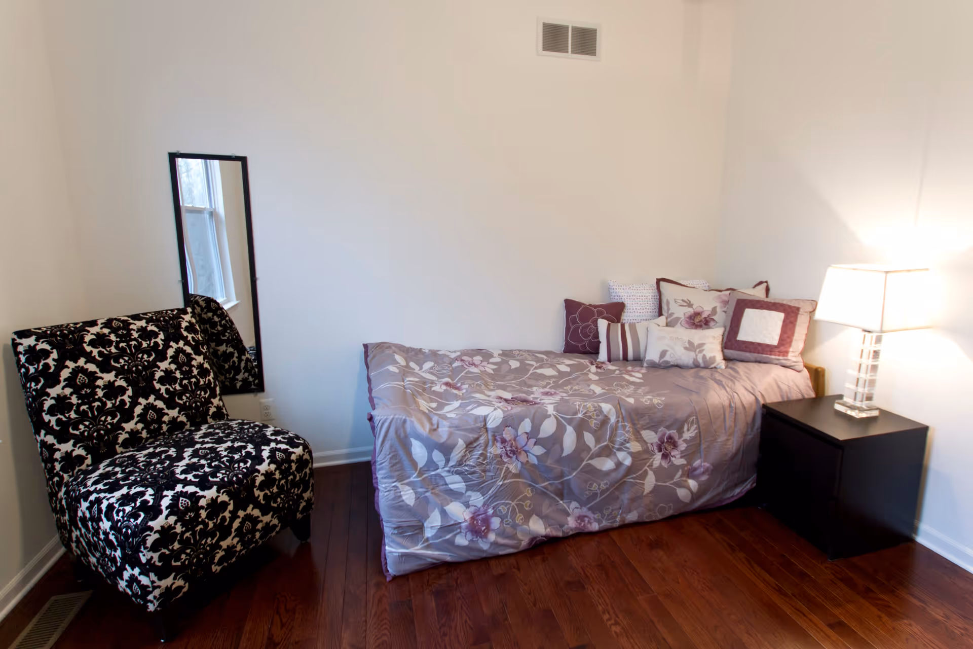 A small bedroom with a single bed covered in a floral patterned comforter and multiple decorative pillows. Next to the bed is a black nightstand with a modern lamp. A black and white patterned armchair is placed near a window with a tall mirror mounted on the wall beside it. The room has wooden flooring and white walls.