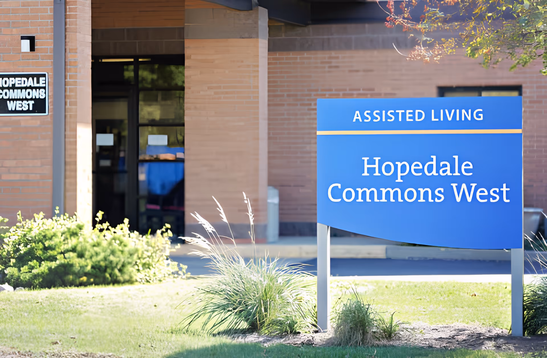 Exterior view of the entrance to Hopedale Commons West Assisted Living facility with a blue sign displaying the facility name and the words 'Assisted Living'. The building is made of brick and there are some green bushes and grass in front.