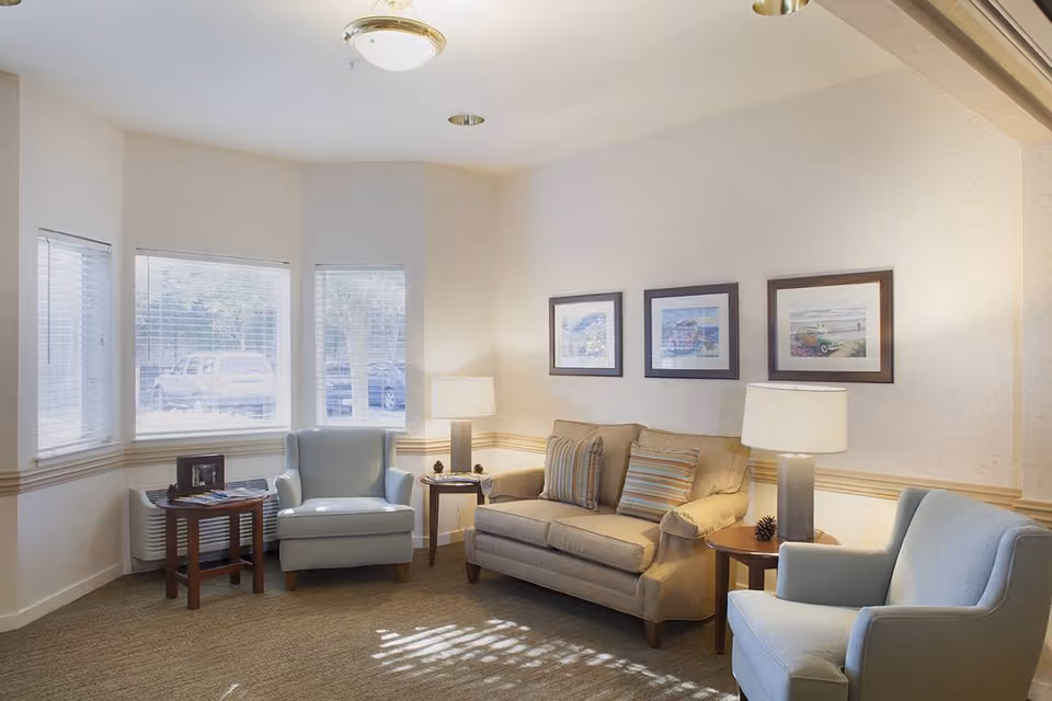 A cozy living room with a beige sofa adorned with striped pillows, two light blue armchairs, two side tables each with a lamp, and three framed pictures on the wall. Large windows allow natural light to fill the room.