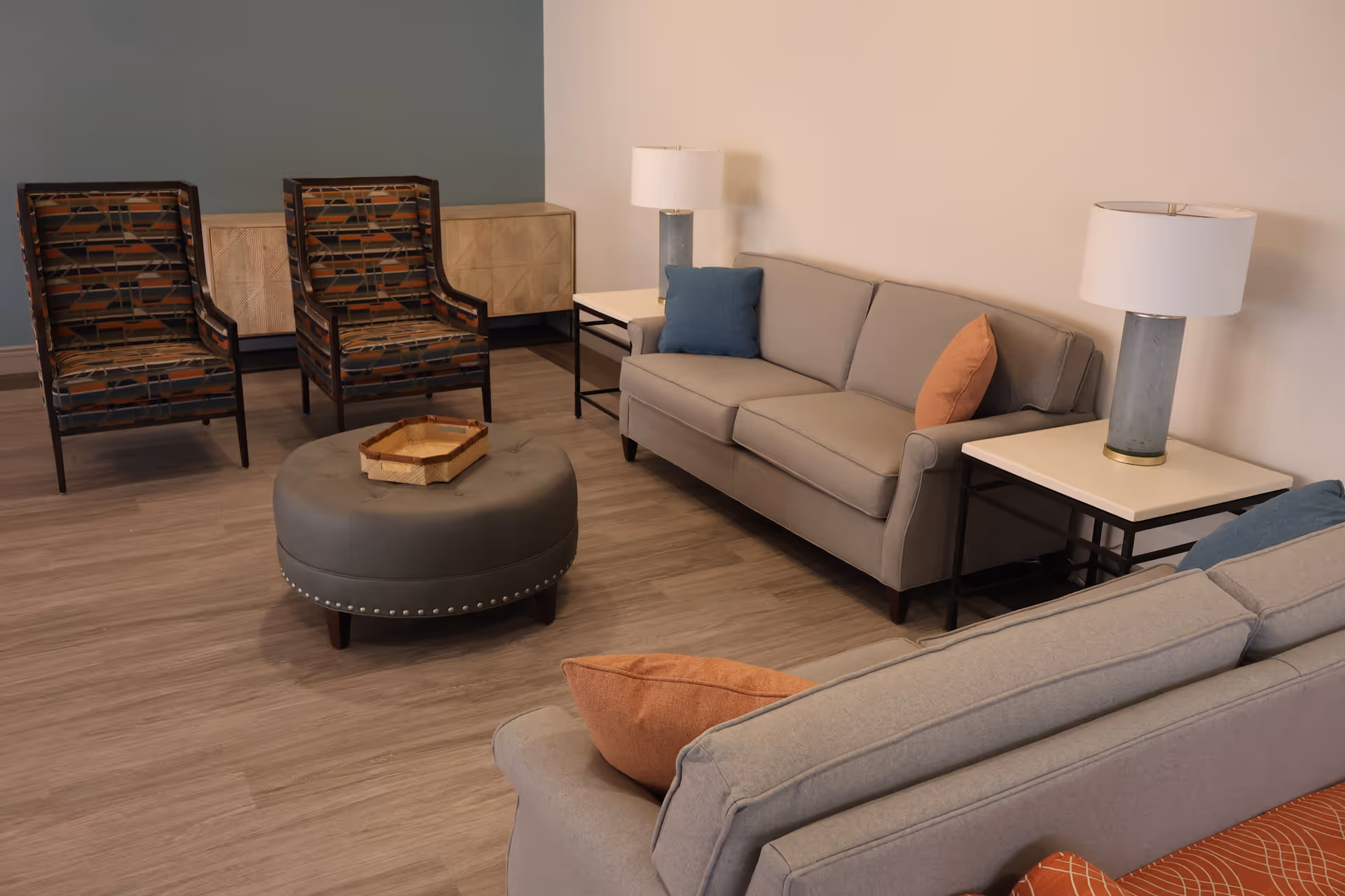 A cozy living room area with two gray sofas adorned with orange and blue pillows, two patterned armchairs, a round gray ottoman with a wooden tray on top, two side tables each with a lamp, and a light blue accent wall behind the armchairs.