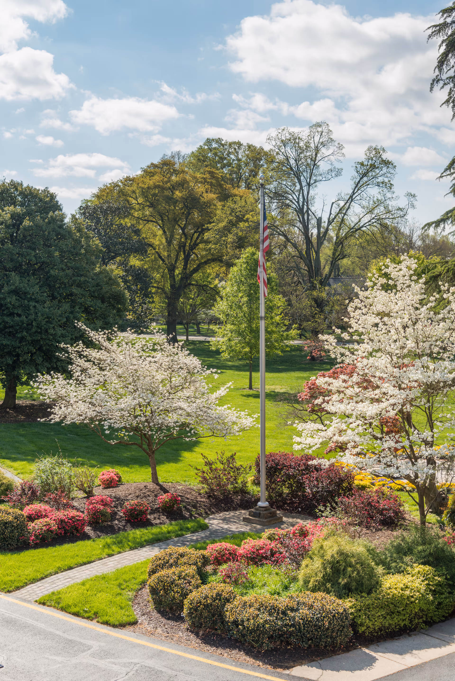 A landscaped garden area with green grass, flowering bushes, and trees in bloom. An American flag on a flagpole stands in the center of the garden. The sky is partly cloudy with blue patches visible.