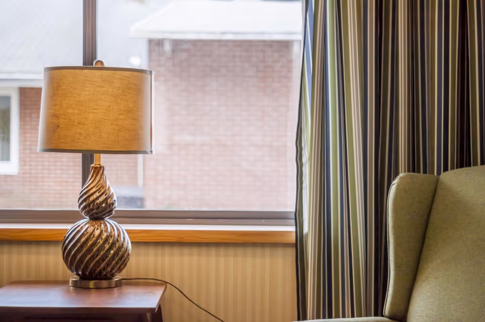 A close-up view of a decorative table lamp with a beige shade on a wooden side table next to a window with striped curtains. Part of a green upholstered armchair is visible on the right side.