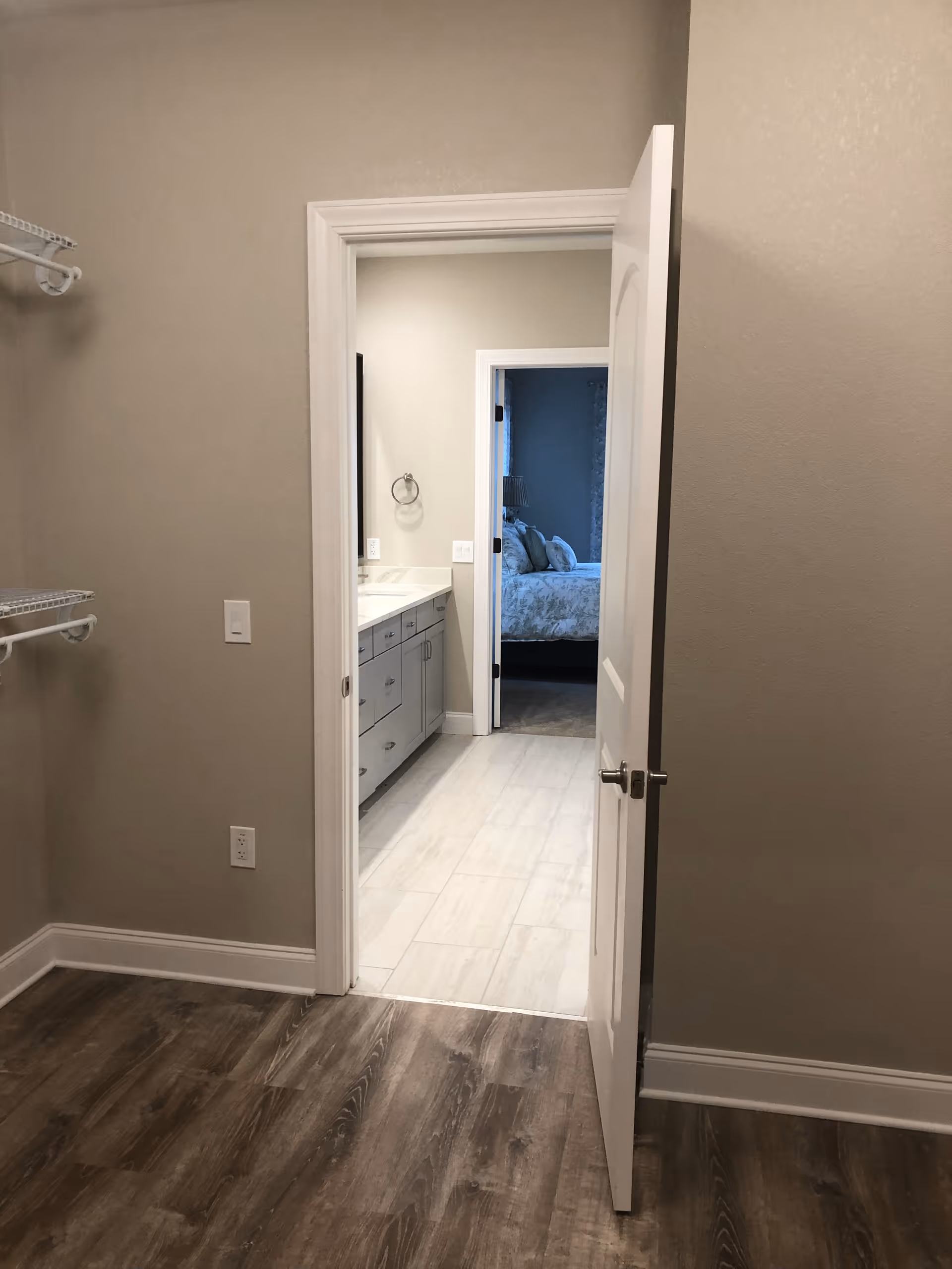 View through an open door from a room with wood flooring and beige walls into a bathroom with white tile flooring, a vanity with drawers and a sink, and a towel ring on the wall. Beyond the bathroom, a bedroom with a bed and pillows is visible.