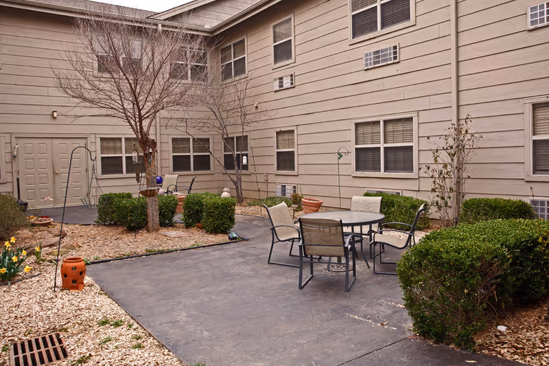 Outdoor courtyard area with a round table and four chairs on a paved surface, surrounded by bushes, leafless trees, and a beige building with multiple windows and air conditioning units.