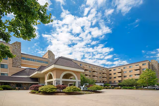 Exterior view of Garden Plaza at Cleveland, a multi-story senior living facility with beige walls and stone accents under a partly cloudy blue sky. The entrance features a covered driveway with arches and landscaped bushes around it.