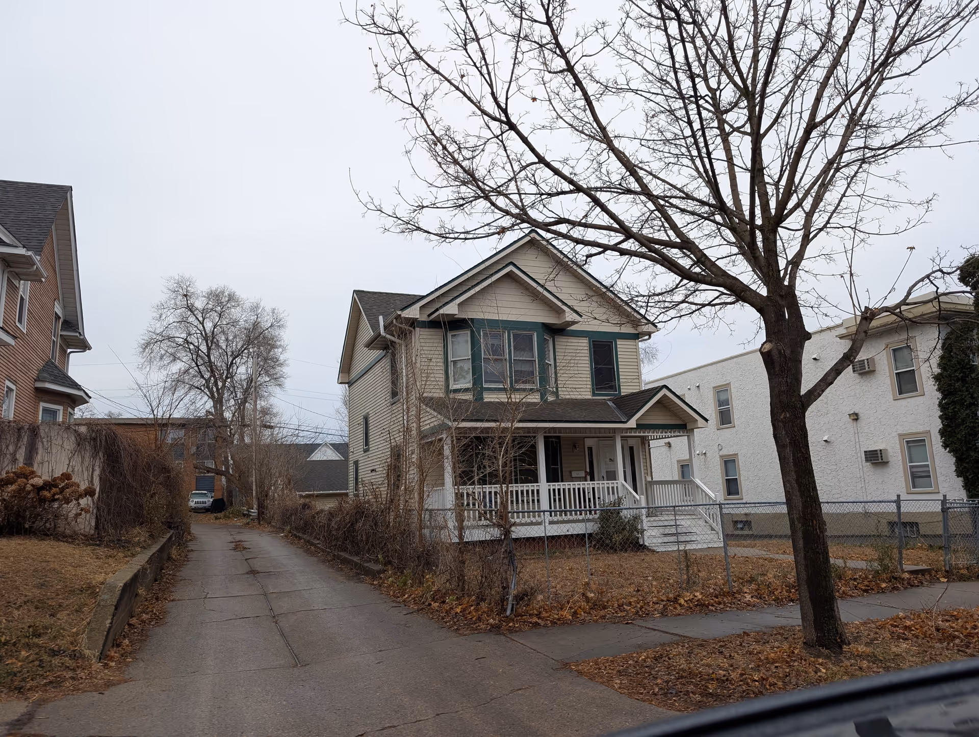 A two-story beige house with green window trim and a front porch with white railings, situated next to a driveway and a leafless tree in the foreground. The sky is overcast and there are neighboring buildings visible on either side.