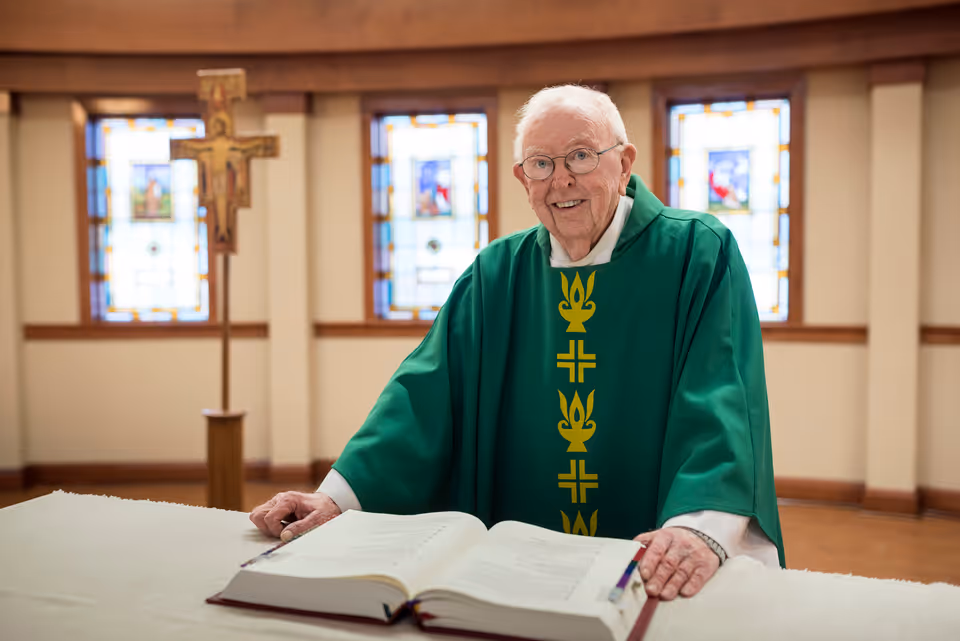An elderly man wearing green religious vestments stands behind a white altar with an open book, smiling. Behind him are three stained glass windows and a wooden crucifix on a stand.
