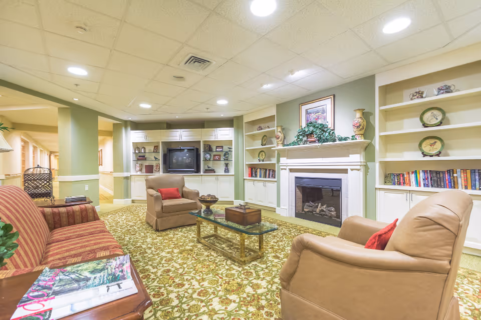 Cozy common lounge with upholstered chairs and a sofa arranged around a glass coffee table facing a fireplace and built-in shelving with a TV.