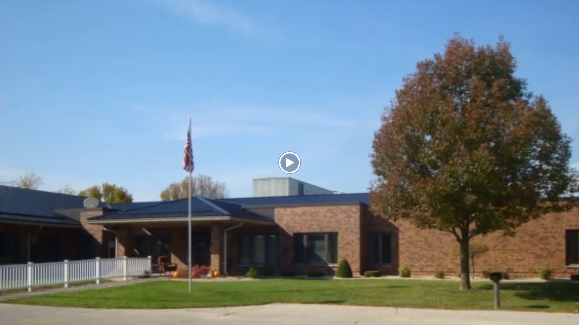 Exterior view of a single-story brick building with a dark roof, a large tree with autumn-colored leaves on the right, a flagpole with an American flag in the center, and a white fence on the left side under a clear blue sky.