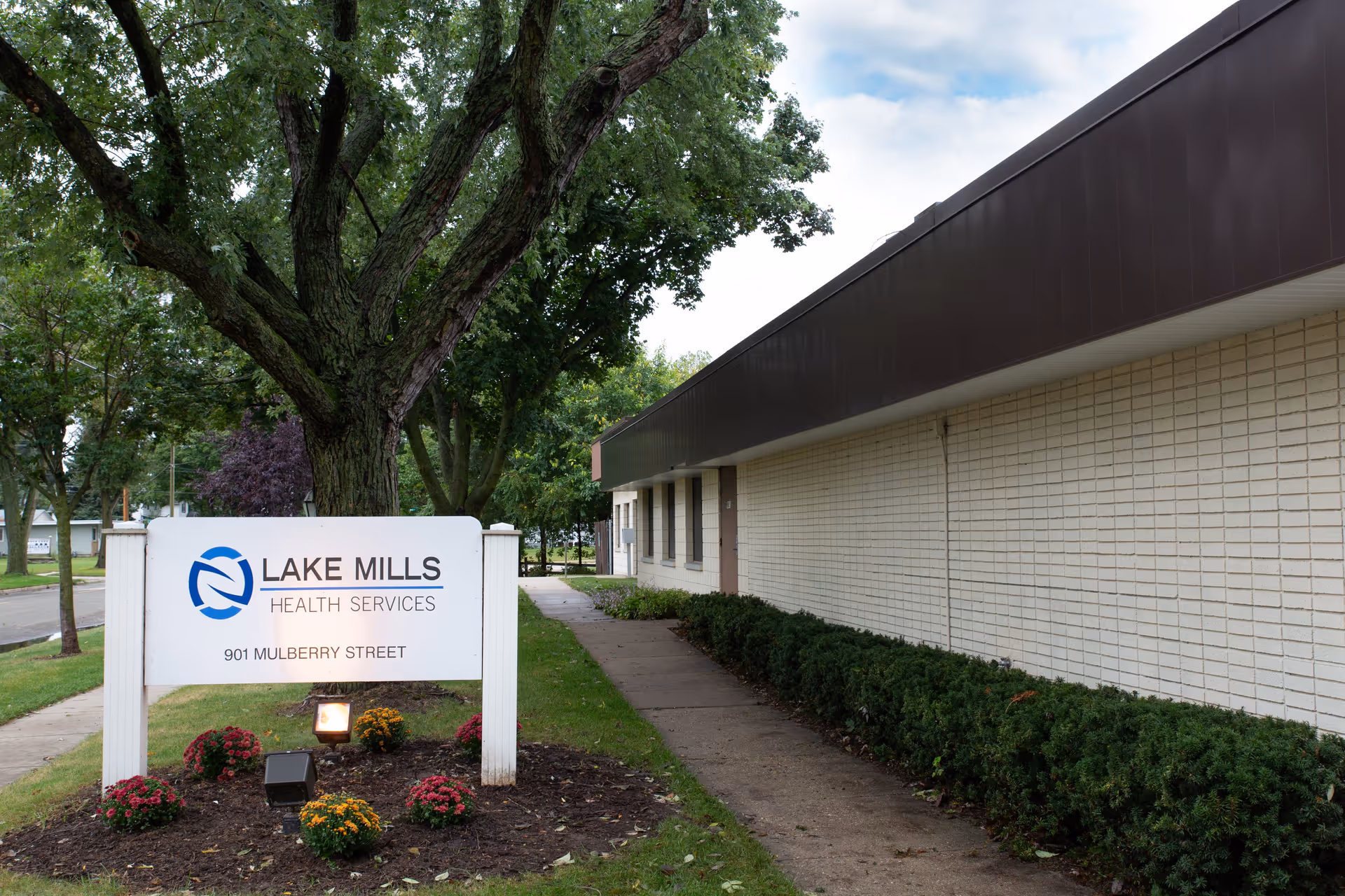 Exterior view of Lake Mills Health Services building with a white sign displaying the facility name and address 901 Mulberry Street, surrounded by trees, bushes, and small flower beds.