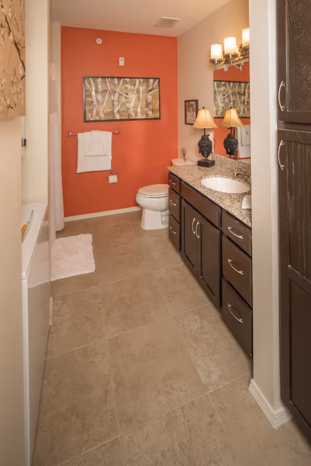 A bathroom with beige tiled floor, a white toilet, a granite countertop with a sink, dark wood cabinets, and a large mirror above the sink. The wall behind the toilet is painted orange and decorated with a framed artwork of birch trees. There is a towel rack with white towels and a small white rug on the floor near the bathtub.