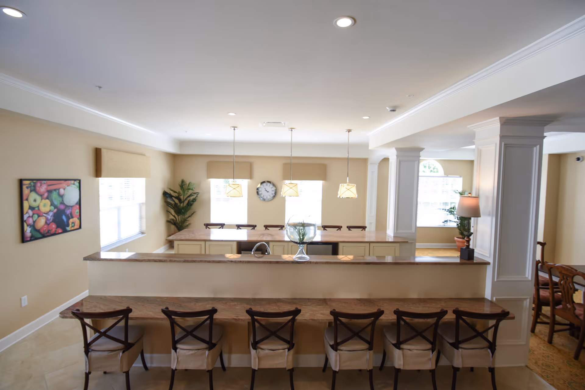 A bright and spacious dining area with a long counter and six chairs in the foreground. Behind the counter is a kitchen island with three pendant lights hanging above it and four chairs. The walls are beige with windows letting in natural light, a clock on the wall, and a framed picture of fruits. There are also some plants and a lamp on a side table near a column.
