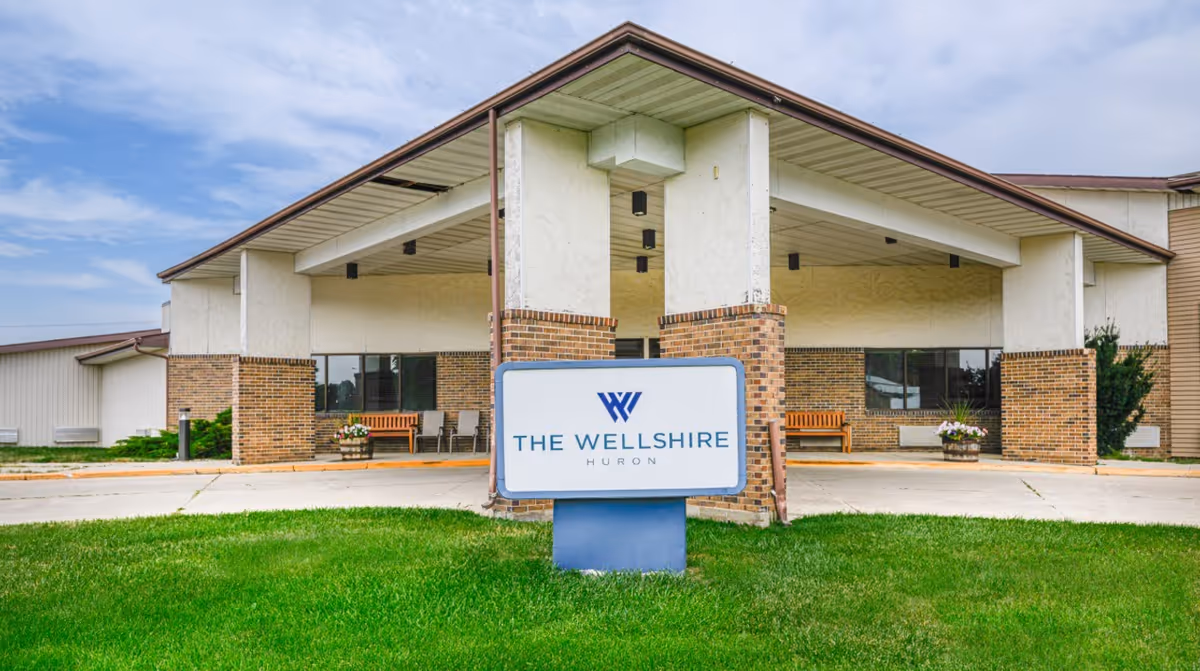 Front exterior view of The Wellshire Huron facility with a covered entrance supported by brick columns, benches on either side, and a sign with the facility's name in front on a grassy area.