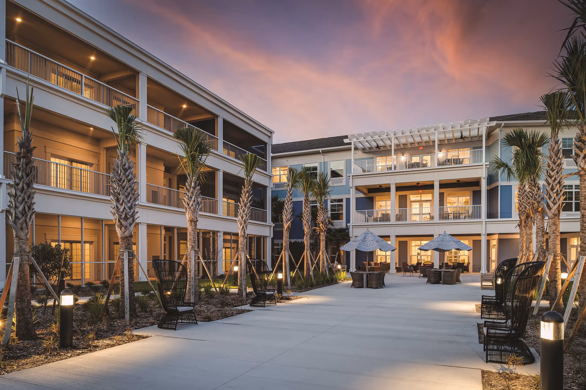 Outdoor courtyard area of a senior living facility at sunset with palm trees lining a wide walkway. The building has three stories with balconies and warm lighting inside. There are outdoor seating areas with tables and umbrellas along the walkway.