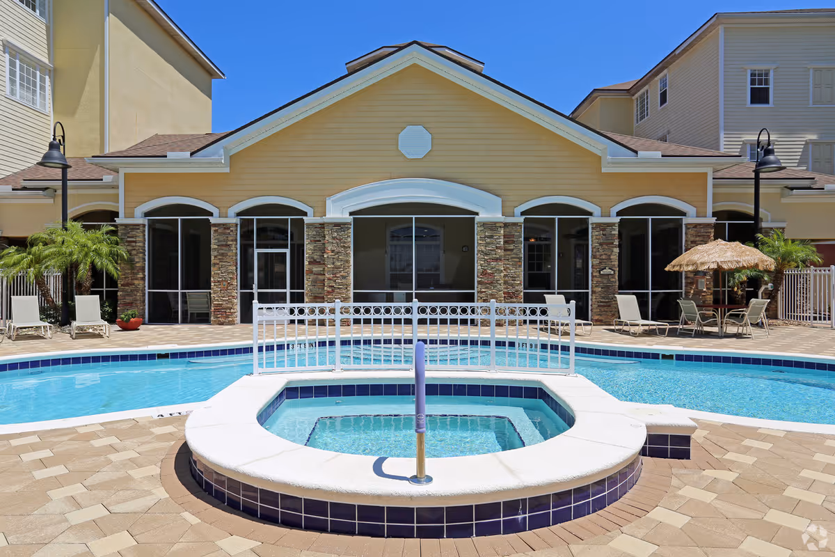 Outdoor swimming pool area with a circular hot tub in the foreground, surrounded by a tiled deck with lounge chairs and a thatched umbrella table. Behind the pool is a yellow building with stone pillars and large windows under a clear blue sky.