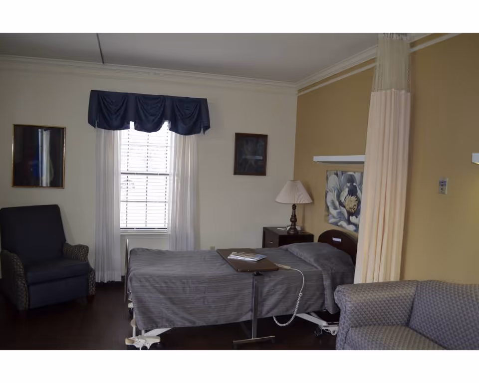 A patient room in a rehabilitation and wellness center featuring a hospital bed with gray bedding, a bedside table with a lamp, a window with white curtains and a blue valance, a dark armchair, a patterned sofa, and a privacy curtain partially drawn.