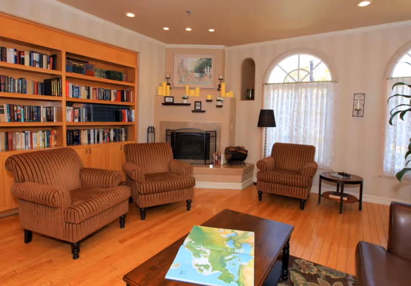 A cozy living room with three striped armchairs arranged around a dark wooden coffee table with a colorful map on it. There is a built-in wooden bookshelf filled with books on the left wall, a fireplace with candles and small framed pictures above it in the center, and two large arched windows with sheer curtains on the right. A floor lamp and a small round side table with a chessboard are also visible.