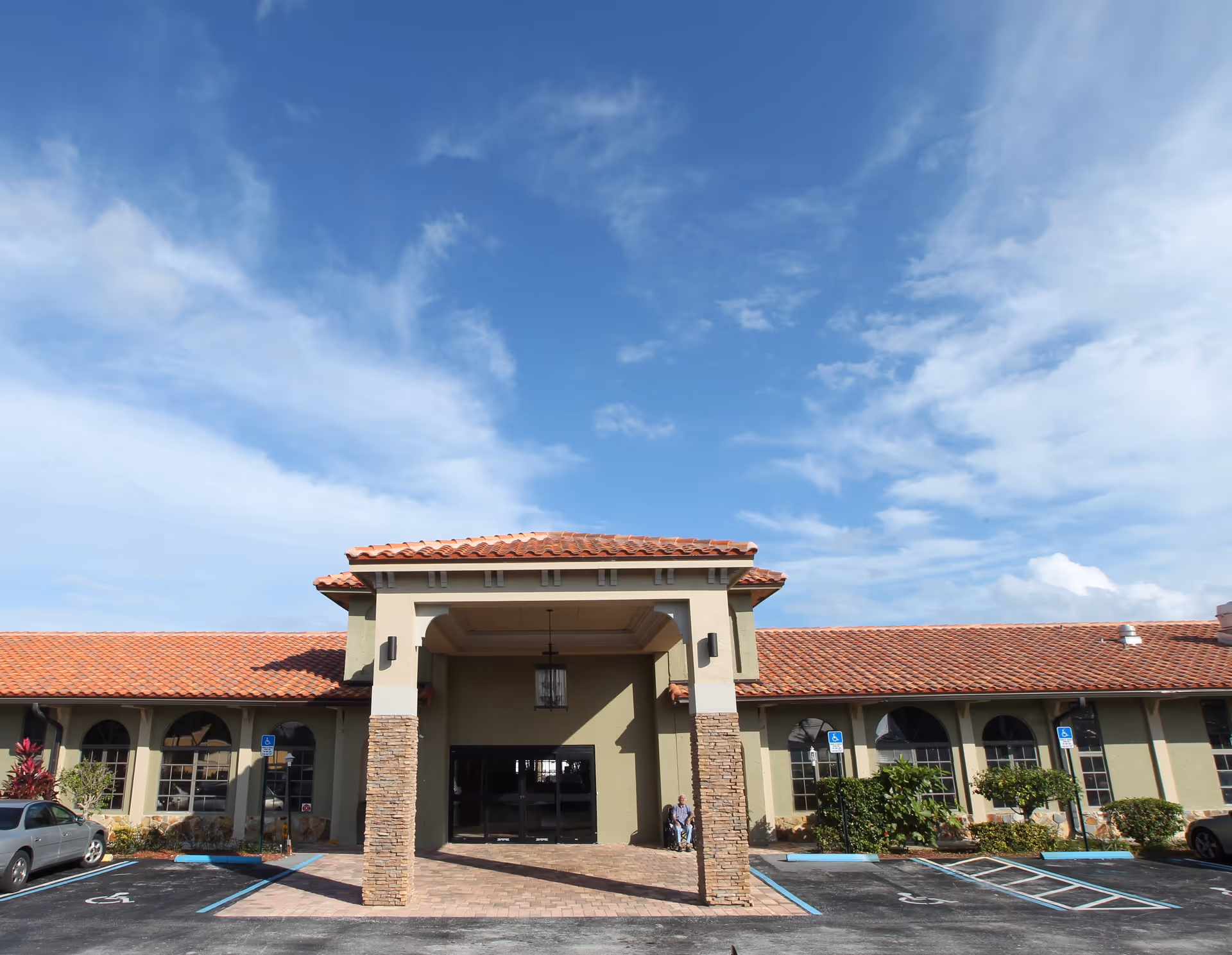 Front exterior view of Boyton Beach Assisted Living facility with a covered entrance supported by stone pillars, a tiled roof, several arched windows, and a parking lot with handicap spaces. A person in a wheelchair is visible near the entrance.
