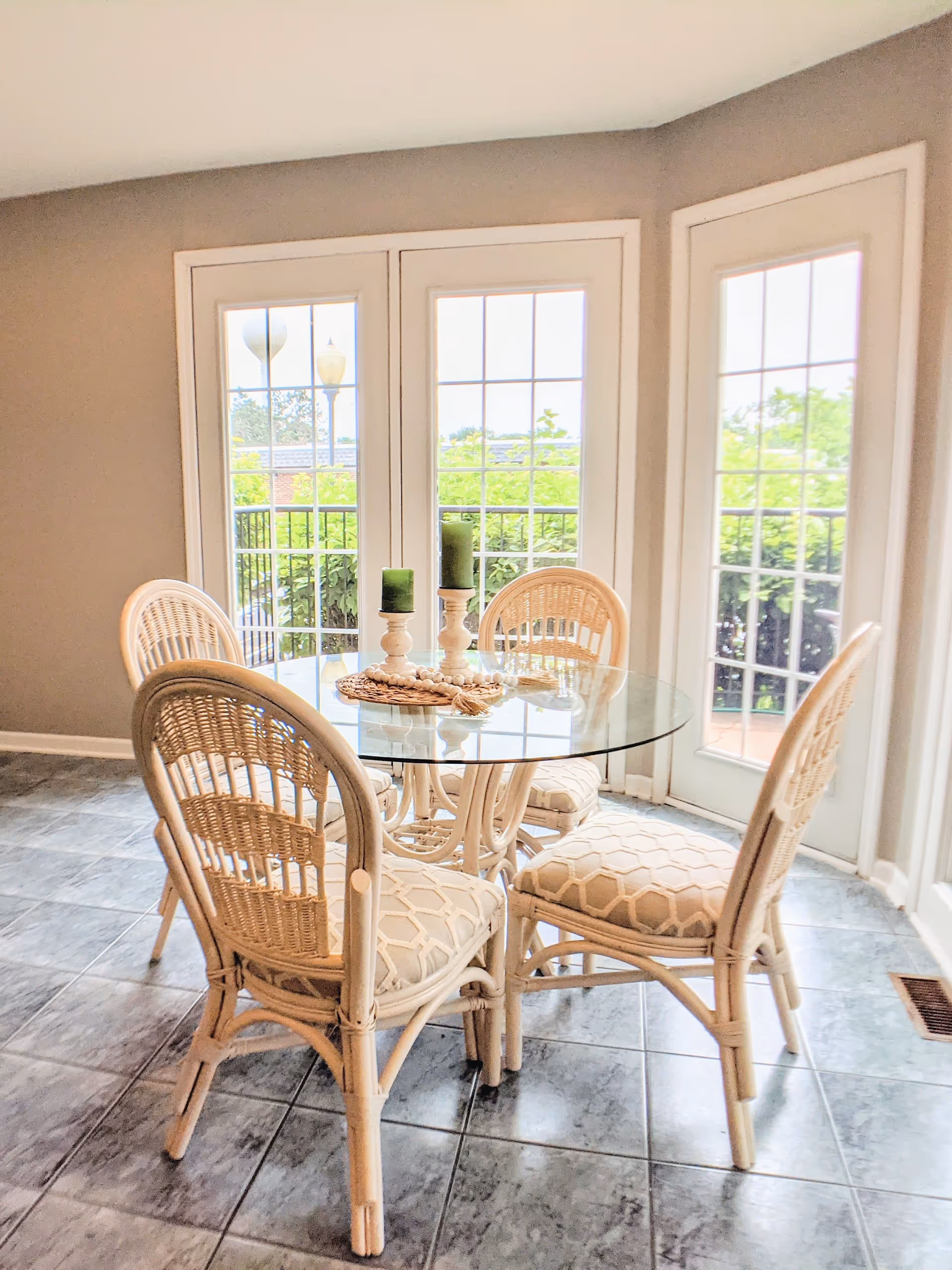 Sunlit dining nook with a round glass table, four wicker chairs and green candles in front of large French doors.