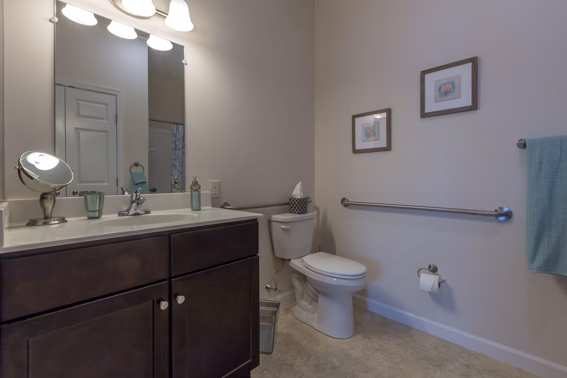 A clean bathroom with a dark wood vanity featuring a white countertop and sink. Above the sink is a large mirror with three light fixtures. To the right is a white toilet with a tissue box on top. The wall has two framed pictures and a metal grab bar. A teal towel hangs on a towel rack on the right side.