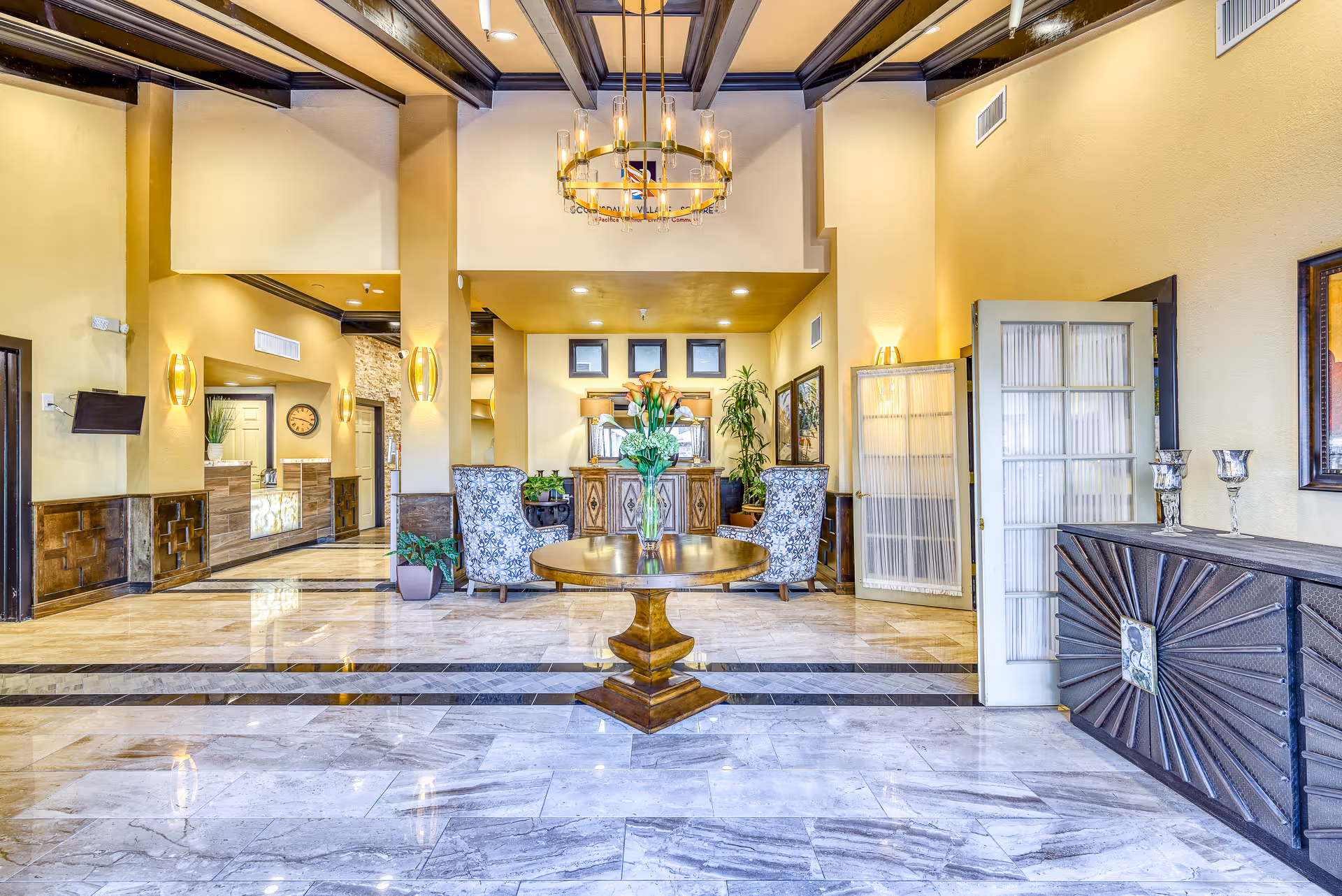 Bright, elegant lobby with marble floors, a central round table with flowers, seating area and reception desk beneath a chandelier.