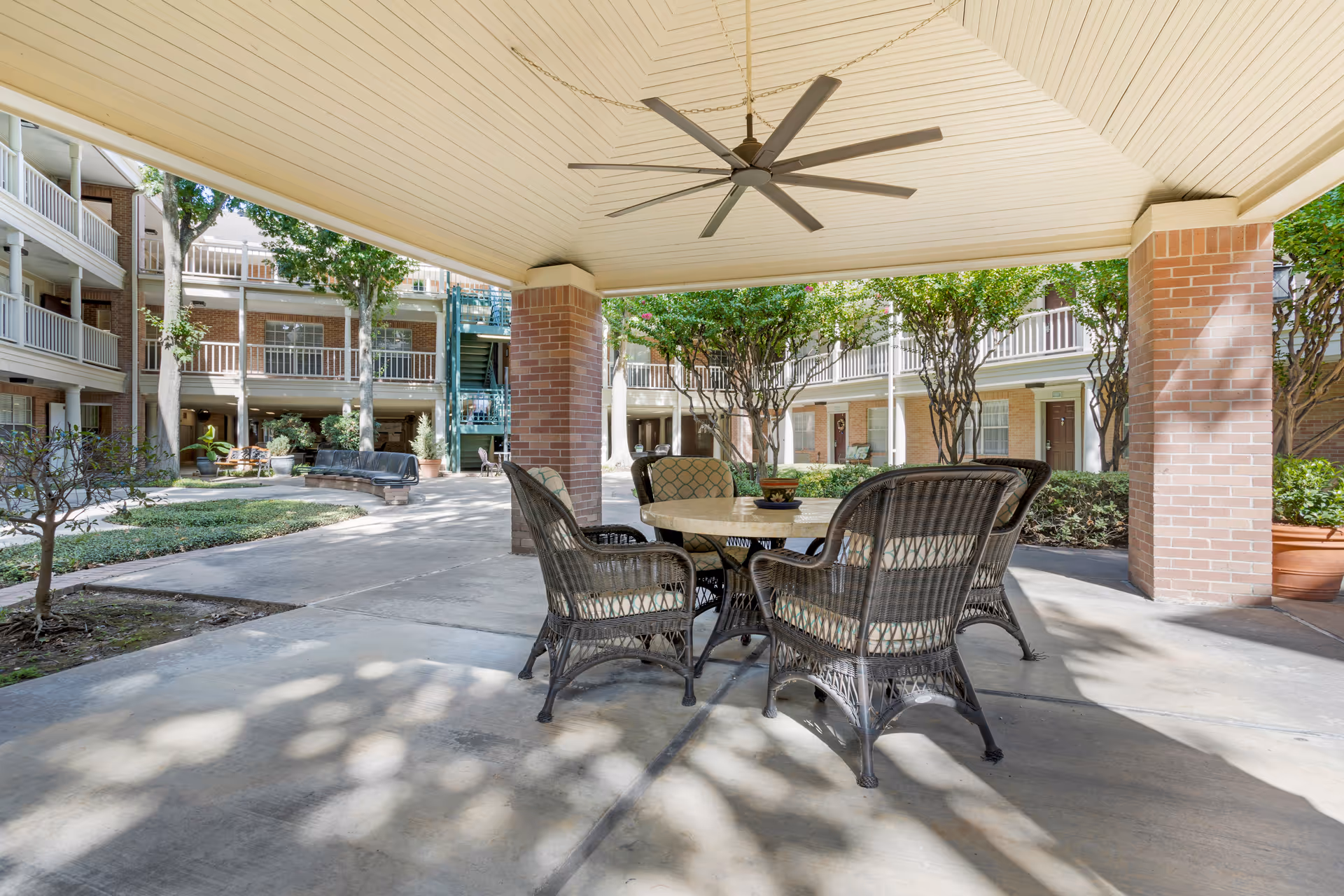 Outdoor covered seating area with a round table and four wicker chairs with cushions, located in the courtyard of a senior living facility. The area is shaded by a roof with a large ceiling fan, surrounded by brick columns, trees, and multi-story building balconies.