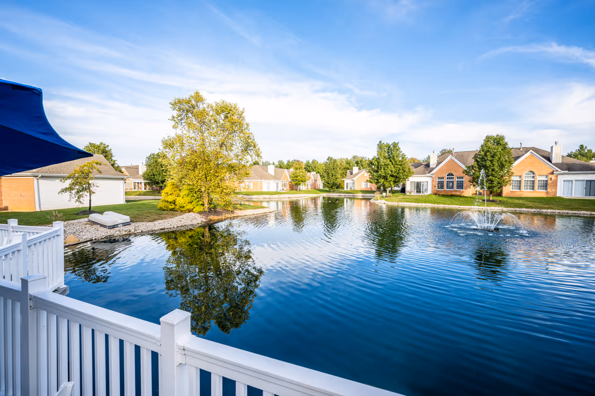 View of a serene pond with a water fountain in the middle, surrounded by green trees and single-story brick buildings under a blue sky with light clouds. A white fence and a blue umbrella are visible in the foreground.