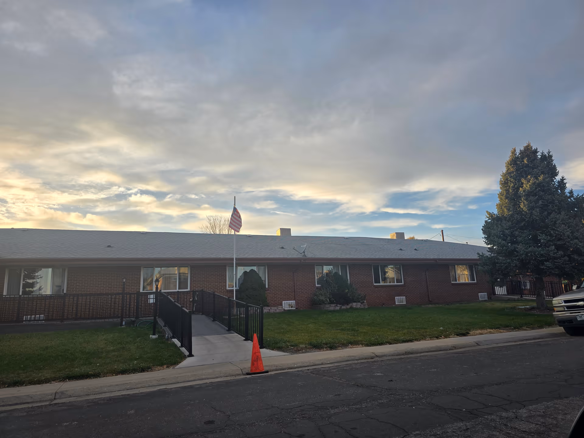 Exterior view of a single-story brick building with several windows, a ramp with black railings leading to the entrance, an American flag on a flagpole, a large tree on the right side, and a parked vehicle partially visible on the right. The sky is partly cloudy with soft sunlight.