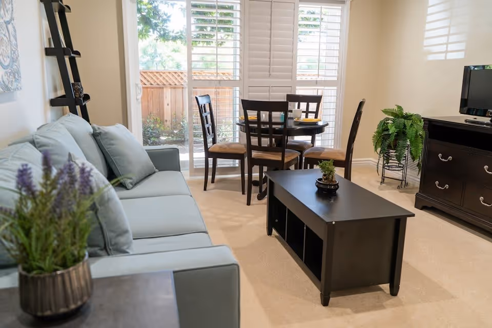 Sunlit living room featuring a light blue sofa, black coffee table, a small dining table with chairs, and sliding glass doors with shutters.