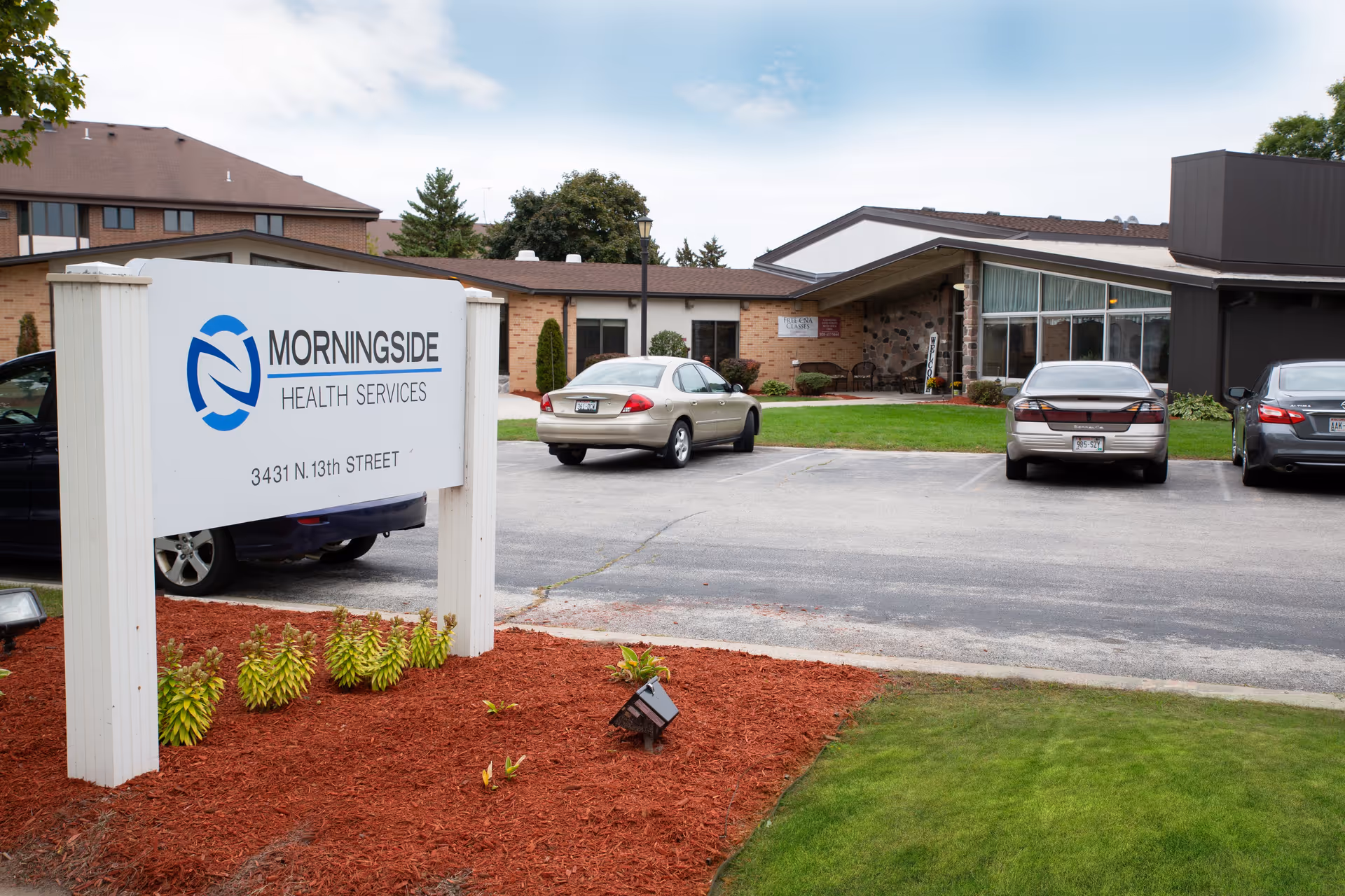 Exterior view of Morningside Health Services building with a parking lot in front. There is a white sign with the facility name and address, some parked cars, and a landscaped area with mulch and small plants.