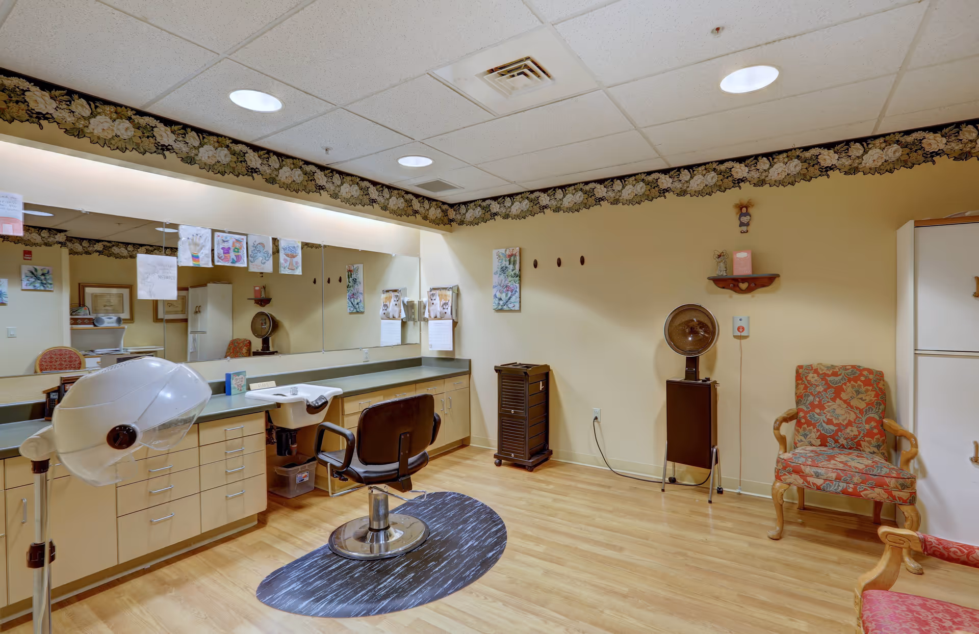 Small salon room with a styling chair, hooded hair dryer, sink and long mirror above cabinets, plus patterned armchairs.