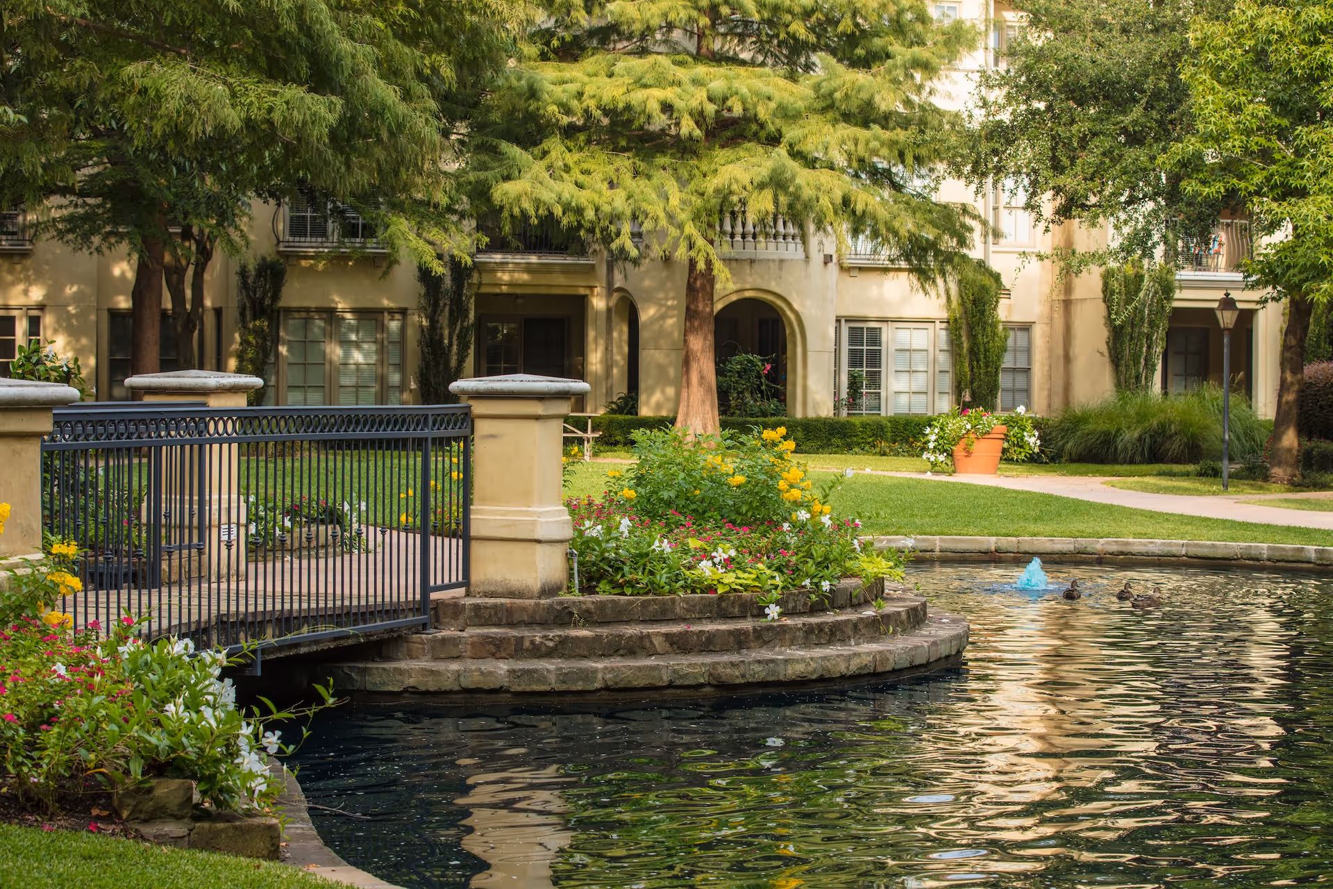 A peaceful outdoor garden area at The Plaza at Edgemere featuring a small pond with a fountain, a stone-bordered flower bed with colorful flowers, a black metal bridge, green grass, trees, and a beige building with windows in the background.