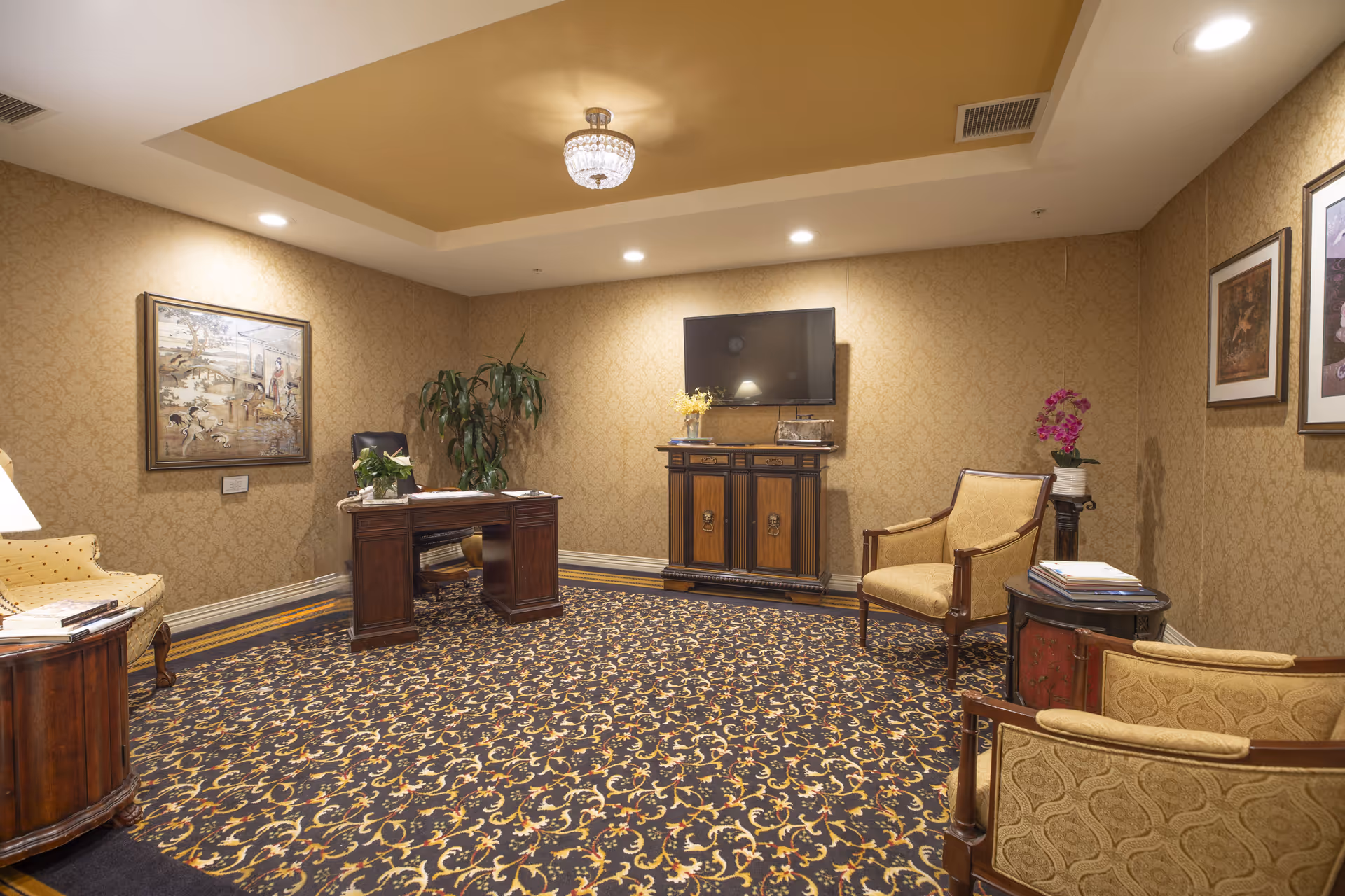 A warmly lit interior room with patterned wallpaper and carpet. The room features a wooden desk with a black chair, a wooden cabinet with a flat-screen TV mounted above it, and several upholstered chairs arranged around the space. There are framed artworks on the walls, a potted plant near the desk, and a small table with a vase of flowers and some magazines.