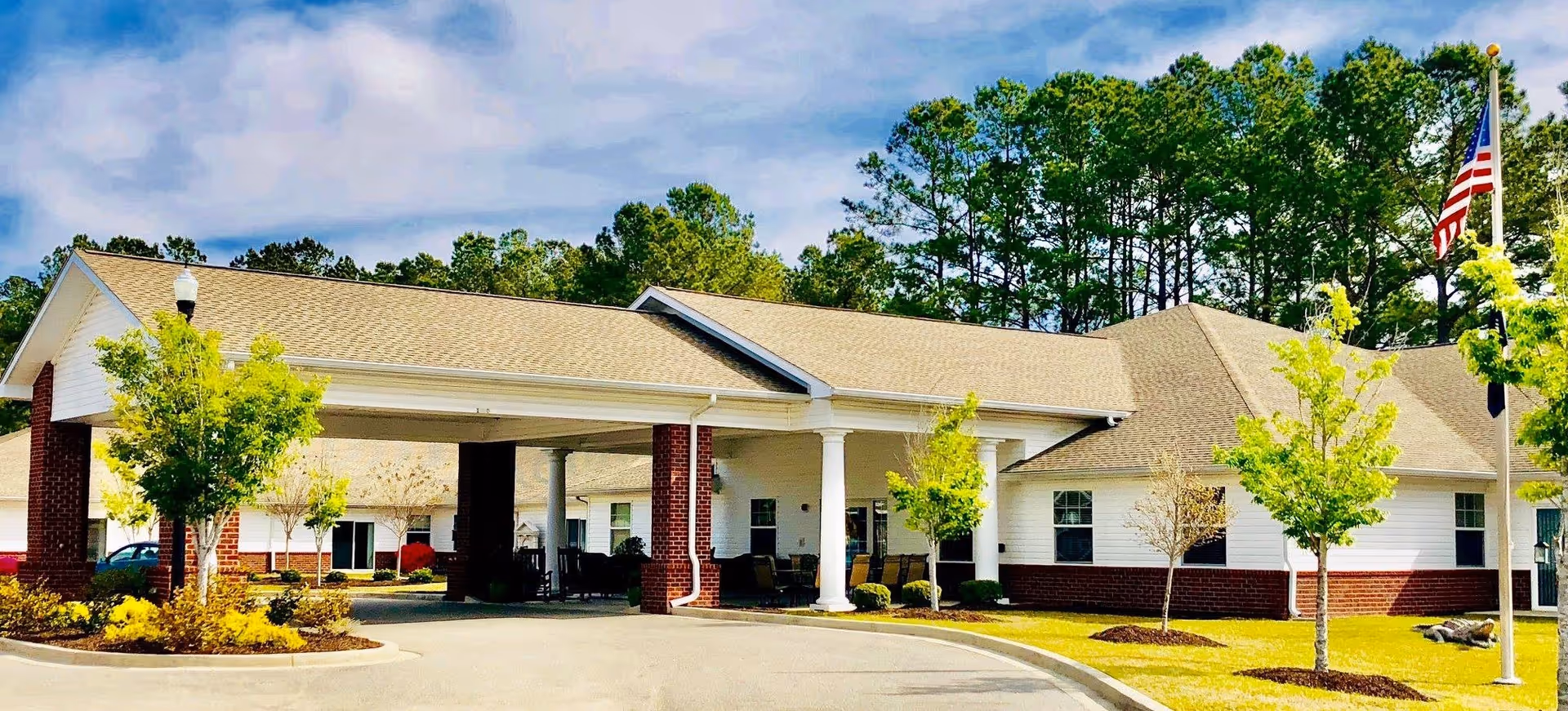 Exterior view of a single-story senior living facility building with a covered entrance supported by white columns. The building has a beige roof, white siding with red brick accents, and is surrounded by small trees and landscaping. An American flag is visible on a flagpole to the right.