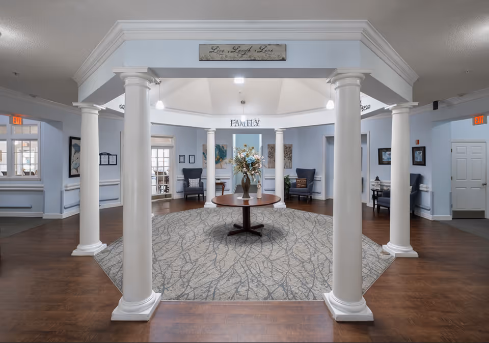 Interior view of a senior living facility lobby area with white columns surrounding a central round table holding a vase of flowers. The walls are painted light blue, and there are several armchairs and framed artwork along the walls. Signs with the words 'Live Laugh Love' and 'FAMILY' are displayed above the columns. The floor is a combination of wood and a patterned carpet.