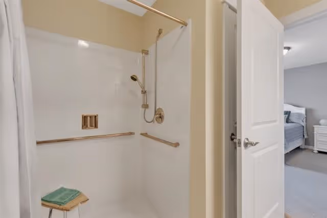 A clean, accessible shower area with grab bars and a handheld showerhead. A small wooden stool with folded green towels is placed inside the shower. The shower is adjacent to a partially open door leading to a bedroom with a bed and nightstand visible.