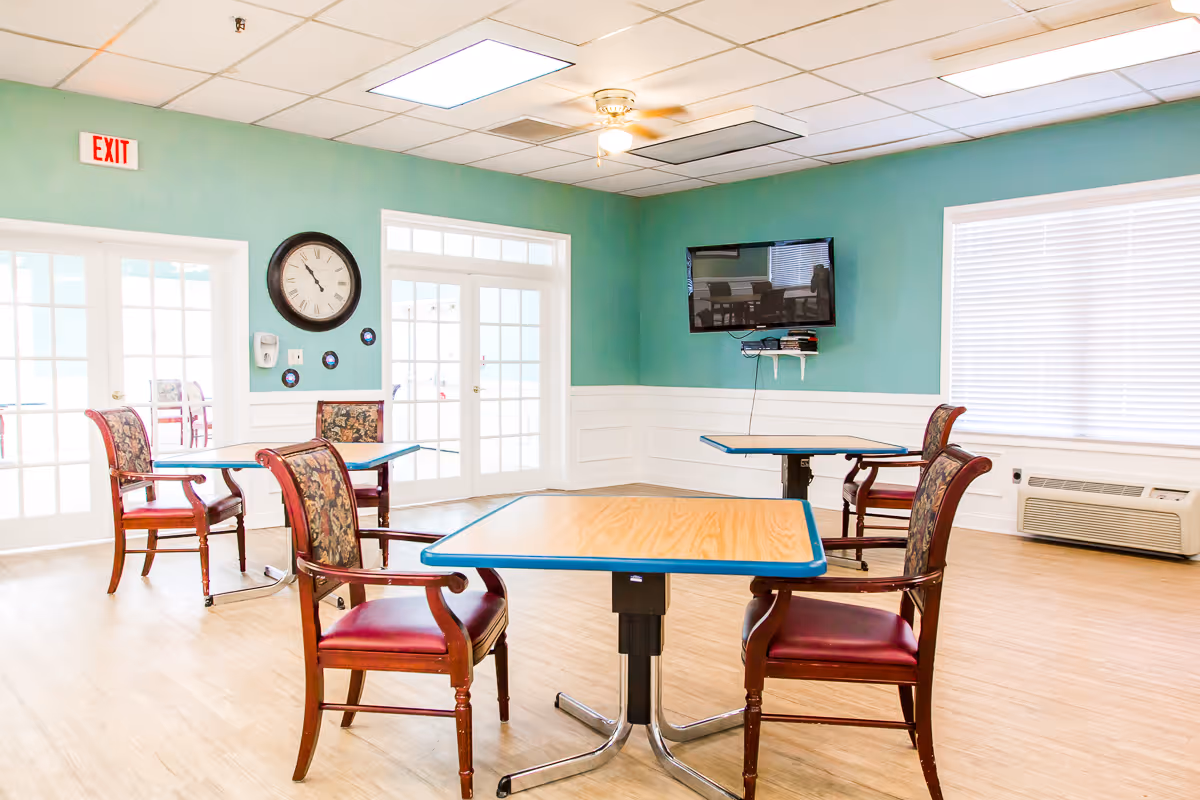 A bright room with light wood flooring and teal walls featuring three tables with wooden chairs that have red cushioned seats and floral backs. A wall clock and a mounted flat-screen TV are visible, along with large windows with blinds and a glass door with an exit sign above it.