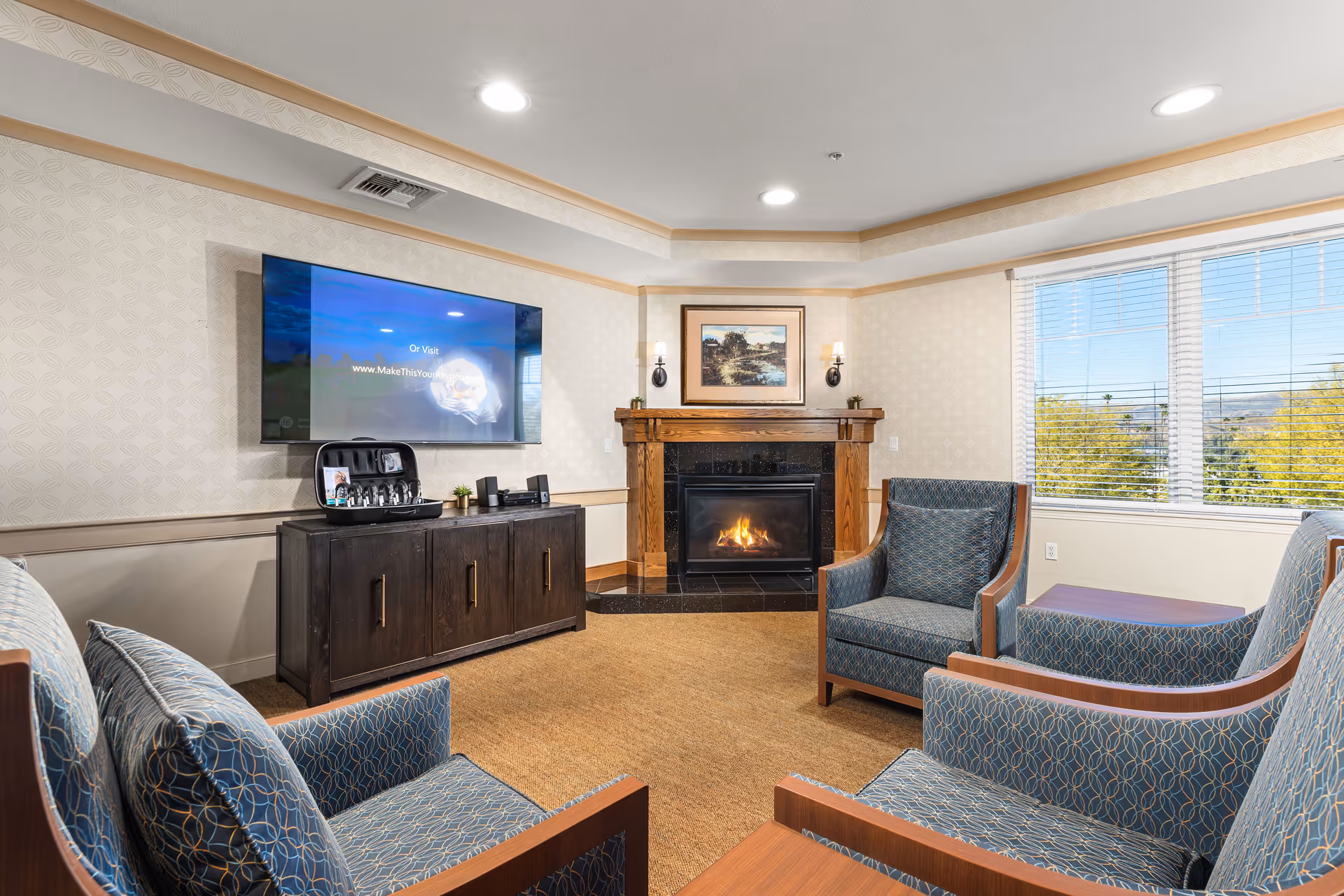 A bright common room with upholstered armchairs facing a fireplace and a wall-mounted TV next to a large window.