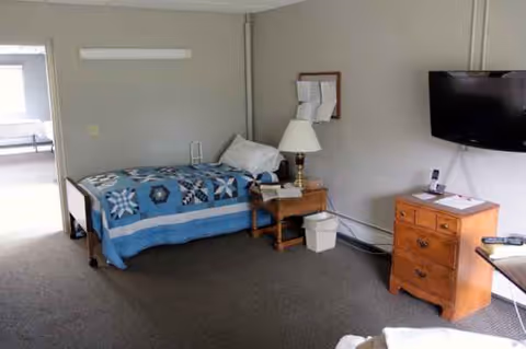 A bedroom in a senior living facility with a bed, dresser, lamp, and wall-mounted TV.