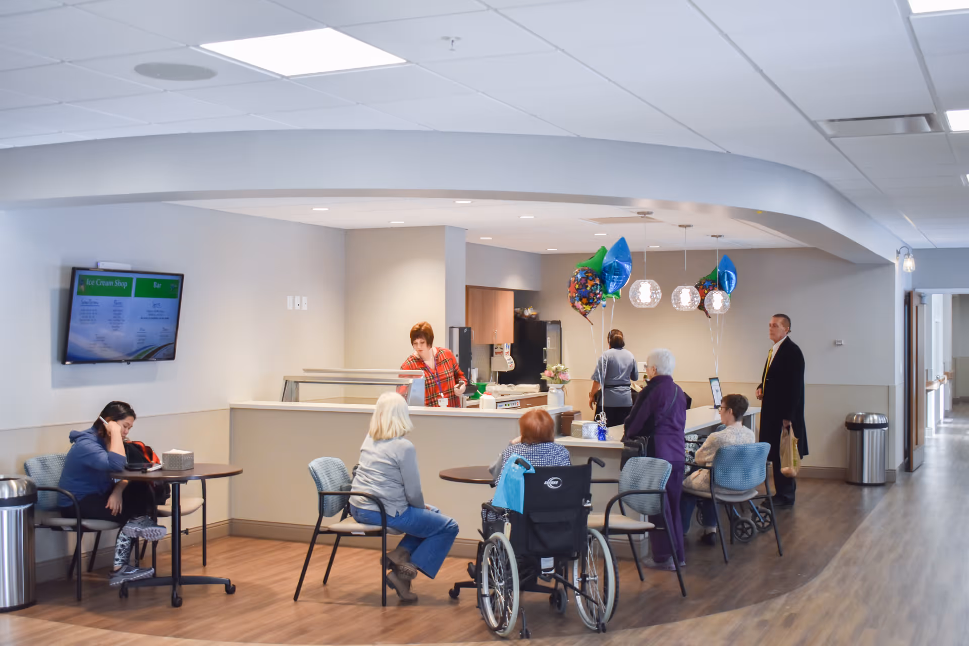 A communal area in a senior living facility with several elderly people seated at tables and in wheelchairs. A staff member behind a counter serves them. The room has modern lighting, balloons, and a menu displayed on a wall-mounted screen.