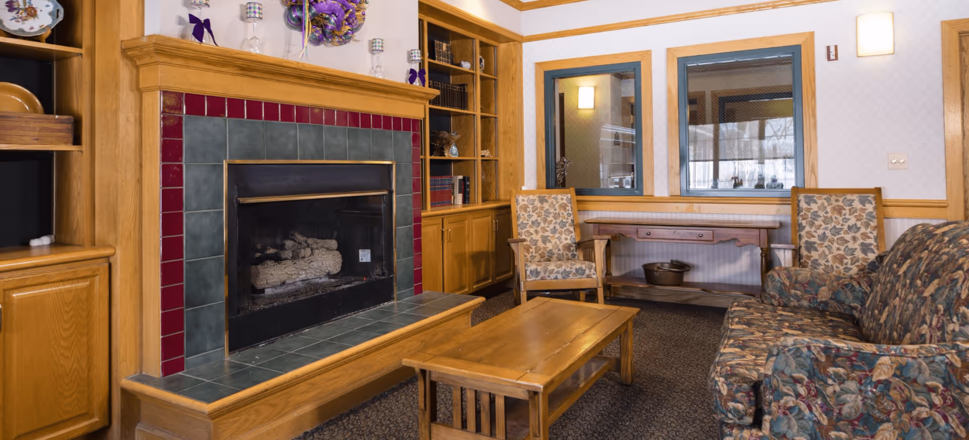 Common room with a tiled fireplace, built-in wood shelving, patterned armchairs and sofa, and a wooden coffee table.