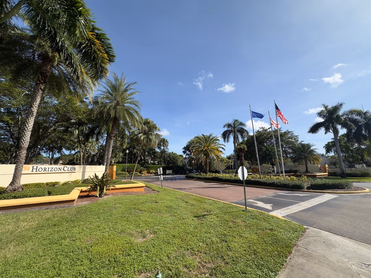Landscaped entrance to The Horizon Club with palm trees, flagpoles, and a driveway under a clear blue sky.