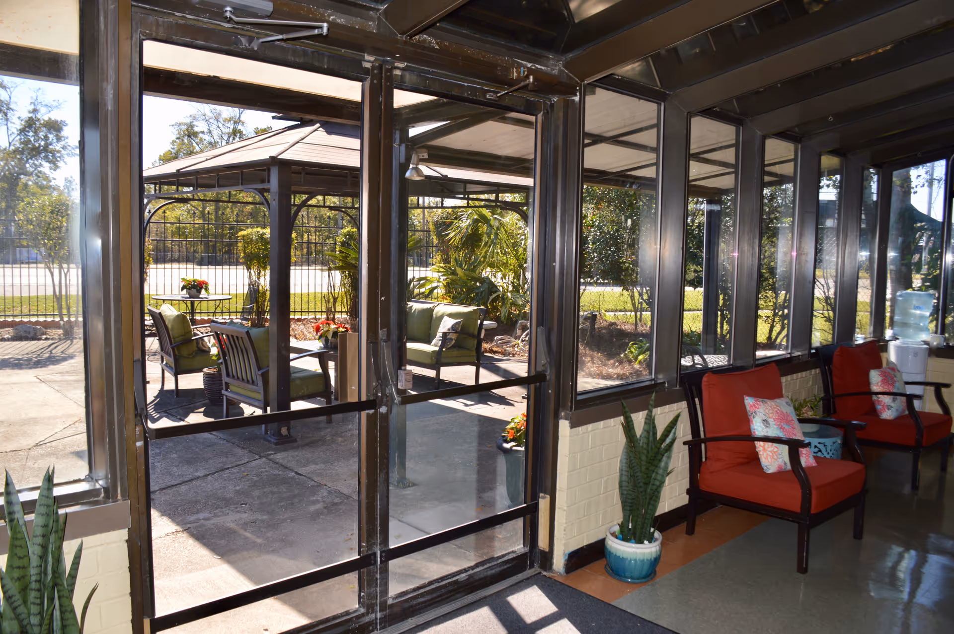 A sunlit indoor seating area with red cushioned chairs and floral pillows next to large windows and a glass door leading to an outdoor patio with green cushioned chairs and a gazebo surrounded by plants and trees.