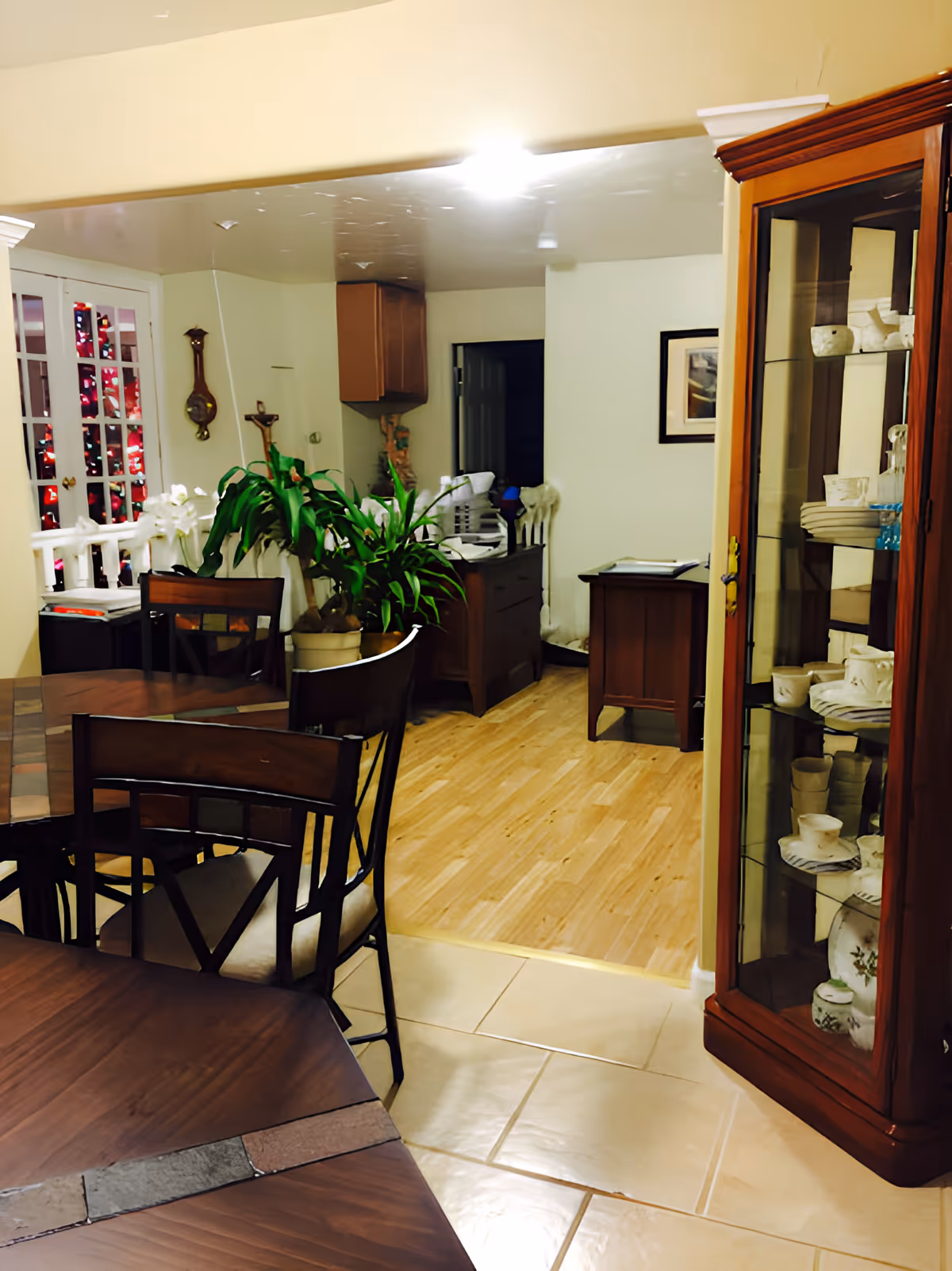 Dining room with a wooden table and chairs, a glass-front china cabinet, potted plants, and a view into an adjacent sitting area.