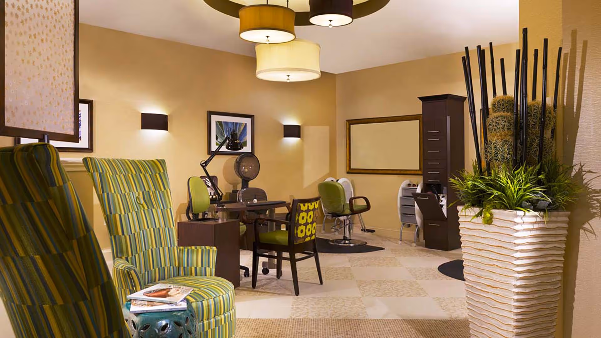 Interior view of a salon area in a senior living facility with striped green armchairs, a manicure table with chairs, hair styling stations with salon chairs, and decorative plants in a large white planter. The walls are beige with framed artwork and modern light fixtures on the ceiling and walls.
