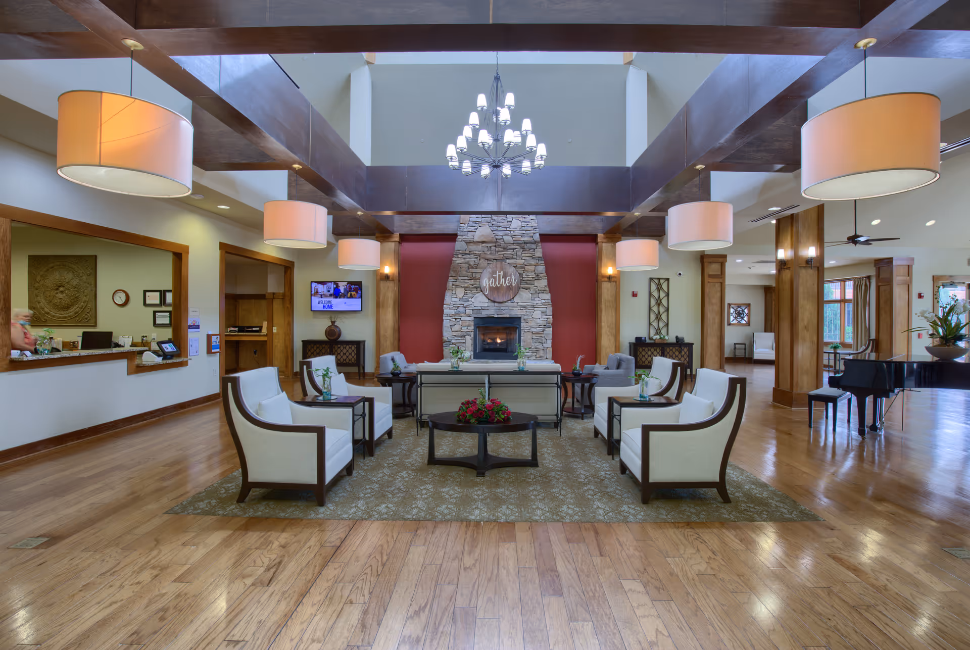 Spacious and well-lit senior living facility common area with wooden floors, a stone fireplace with a 'gather' sign above it, multiple white cushioned chairs arranged around a central coffee table with flowers, large hanging lamps, and a grand piano to the right.