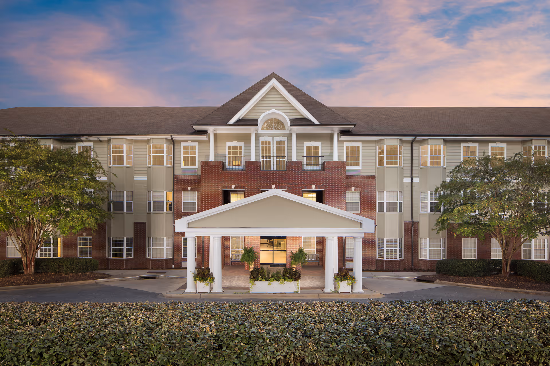 Front entrance of a three-story senior living building with a covered porte-cochere, brick facade, and landscaping under a colorful sky.