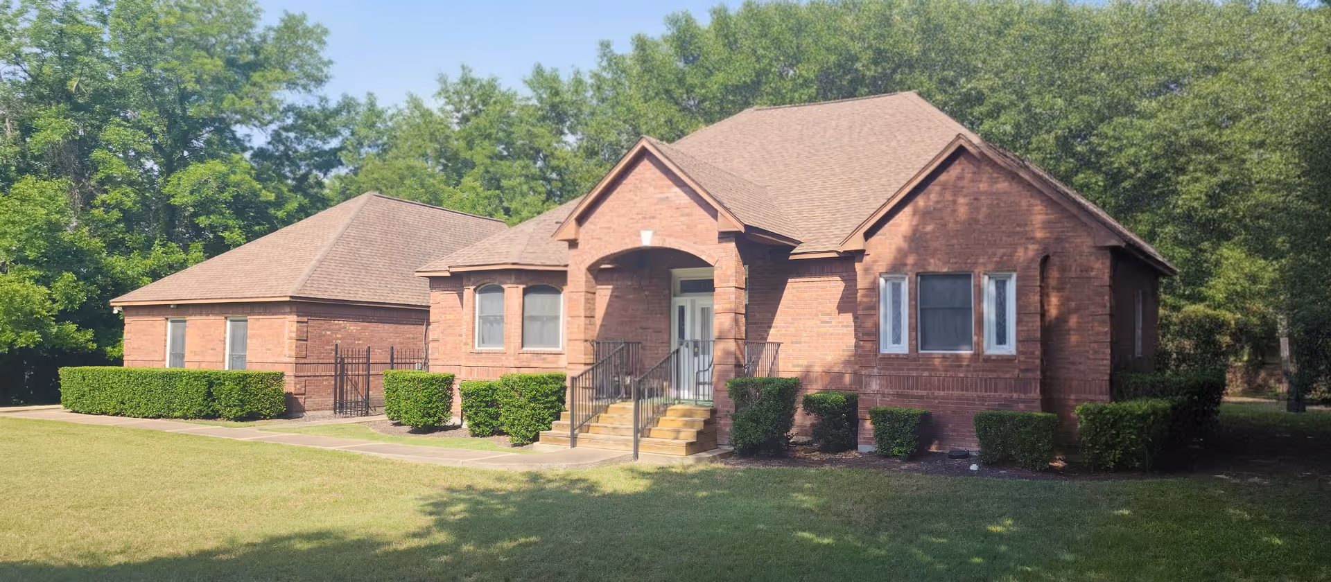 A single-story brick building with a gabled roof, a small front porch with steps and black railings, surrounded by neatly trimmed bushes and a well-maintained lawn, with trees in the background.