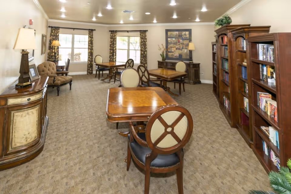 A well-lit room with multiple wooden tables and chairs arranged for seating. The room features large windows with patterned curtains, a carpeted floor, wooden bookshelves filled with books, and decorative lamps on side tables. The overall decor is warm and inviting, suitable for reading or socializing.