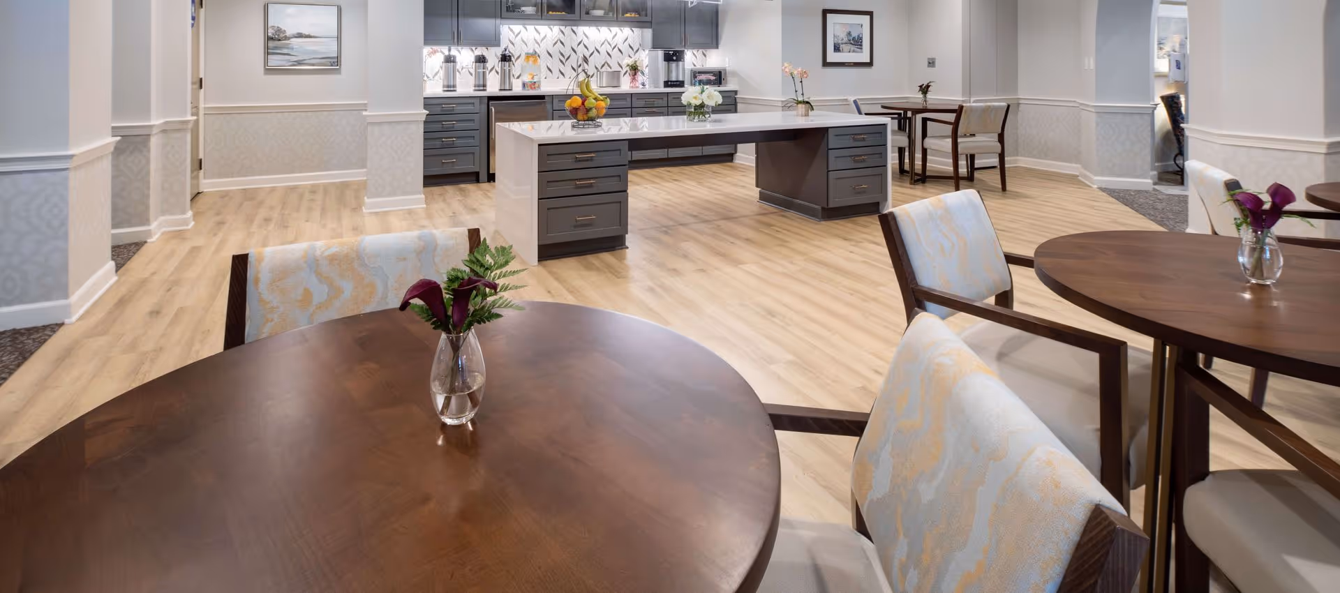 Bright common dining area with round wooden tables and upholstered chairs facing a modern kitchen island.